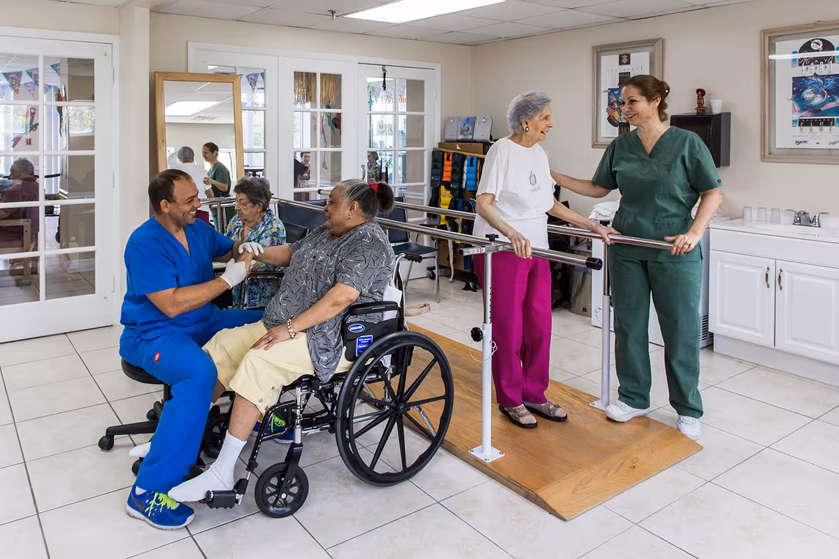 A rehabilitation room in a senior living facility where a healthcare worker in blue scrubs is assisting a woman in a wheelchair, while another healthcare worker in green scrubs helps an elderly woman practice walking using parallel bars. The room has tiled floors, white walls, and framed pictures on the wall.