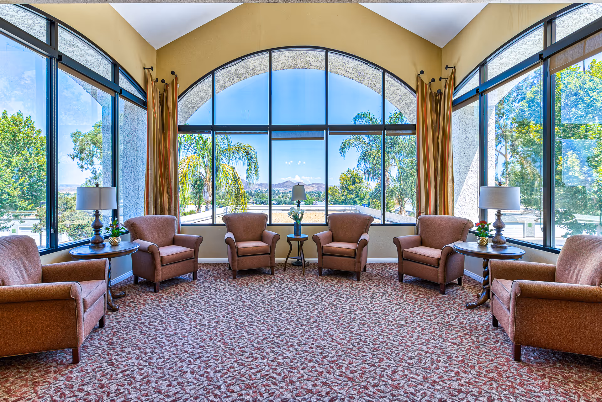 A bright and spacious sitting area with six brown upholstered armchairs arranged in a semi-circle around two small wooden tables with lamps and plants. The room features large floor-to-ceiling windows with an arched design, offering a clear view of palm trees and distant mountains under a blue sky.