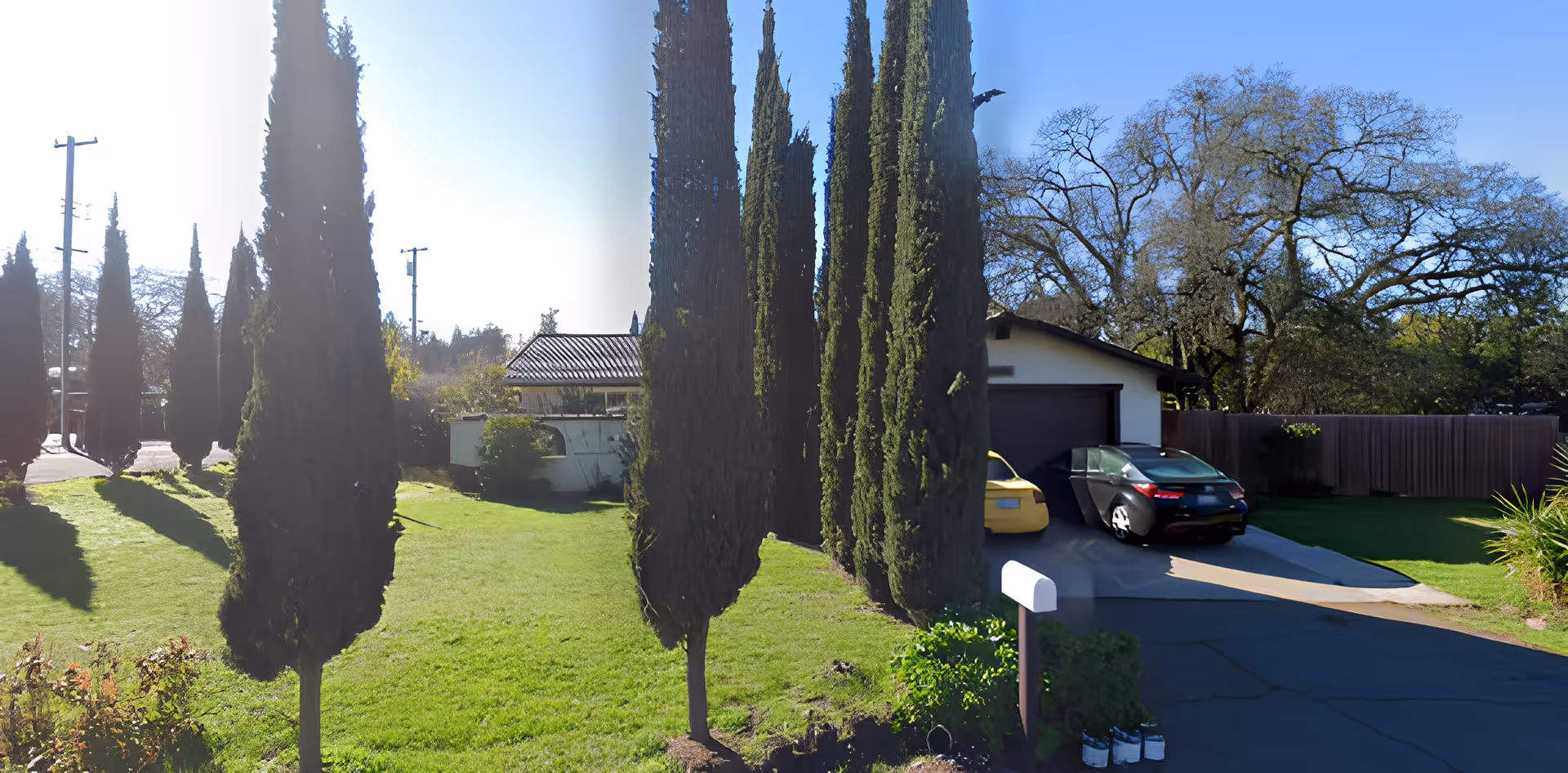Outdoor view of La Serena House showing a green lawn with tall, narrow trees lined up along a pathway. In the background, there is a white building with a dark garage door and two parked cars, one yellow and one black. A mailbox and some plants are visible near the driveway.