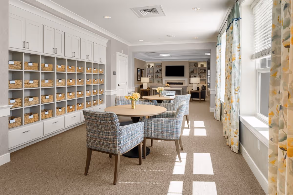 A bright and spacious common area in a senior living facility featuring round tables with plaid upholstered chairs. Along one wall, there are built-in shelves filled with wicker baskets. The room has large windows with floral curtains allowing natural light to fill the space. In the background, there is a seating area with a fireplace, lamps, and a wall-mounted TV.
