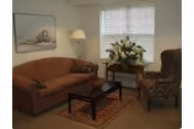 A cozy living room with a brown sofa, a patterned armchair, a wooden coffee table on a decorative rug, a side table with a large floral arrangement, a floor lamp, and a framed picture on the wall.
