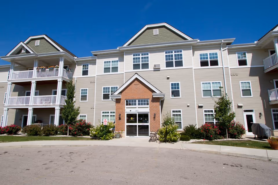 Exterior view of Granville Heights Senior Apartments, a three-story building with beige siding and green gabled roofs. The entrance features a brick facade with glass doors, surrounded by landscaped bushes and small trees. There are balconies on the left and right sides of the building, and a clear blue sky overhead.