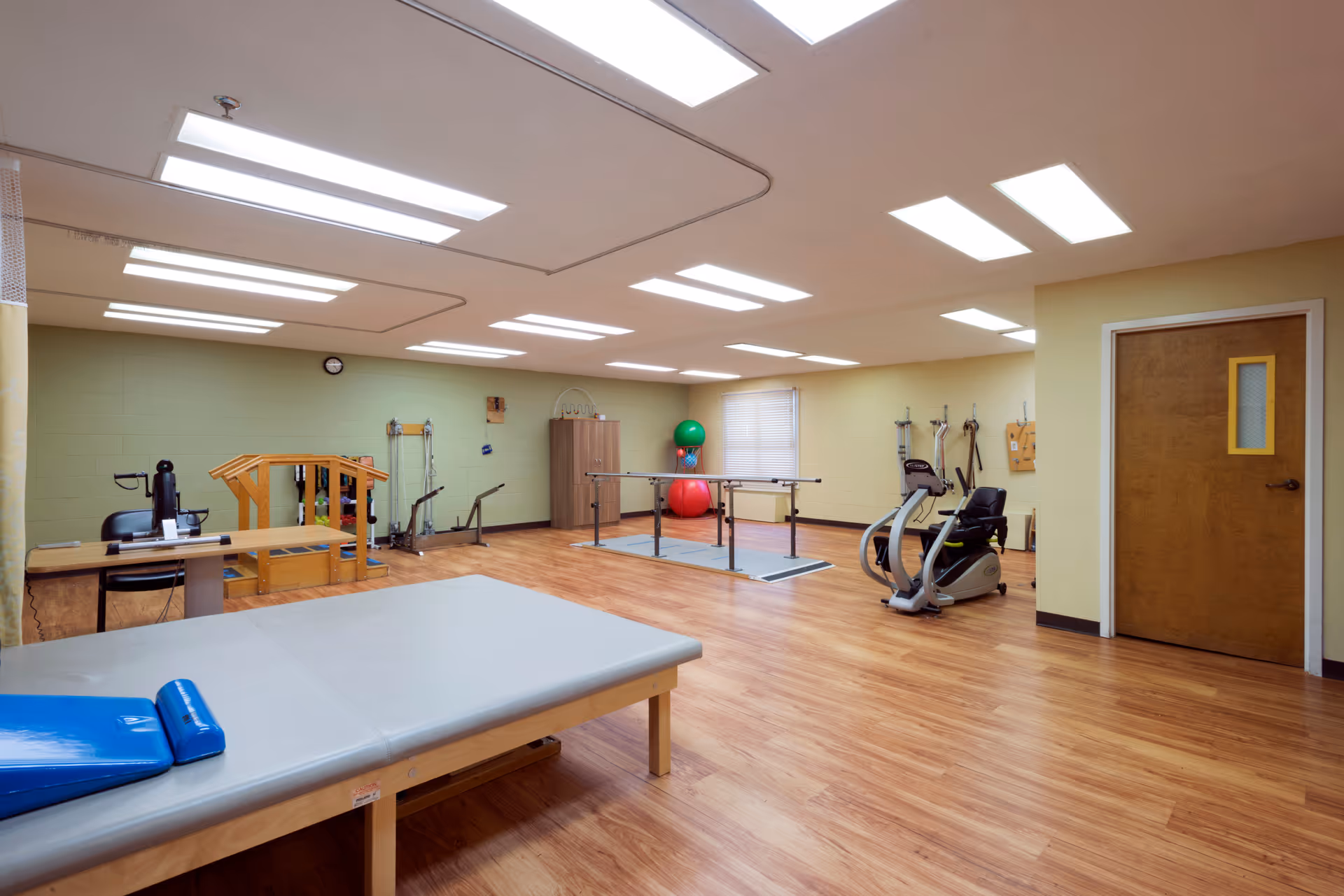 A spacious physical therapy room with wooden flooring and beige walls. The room contains various exercise equipment including parallel bars, exercise balls, a stationary bike, and a therapy table with a blue cushion. There is a wooden door on the right side and a clock on the far wall.