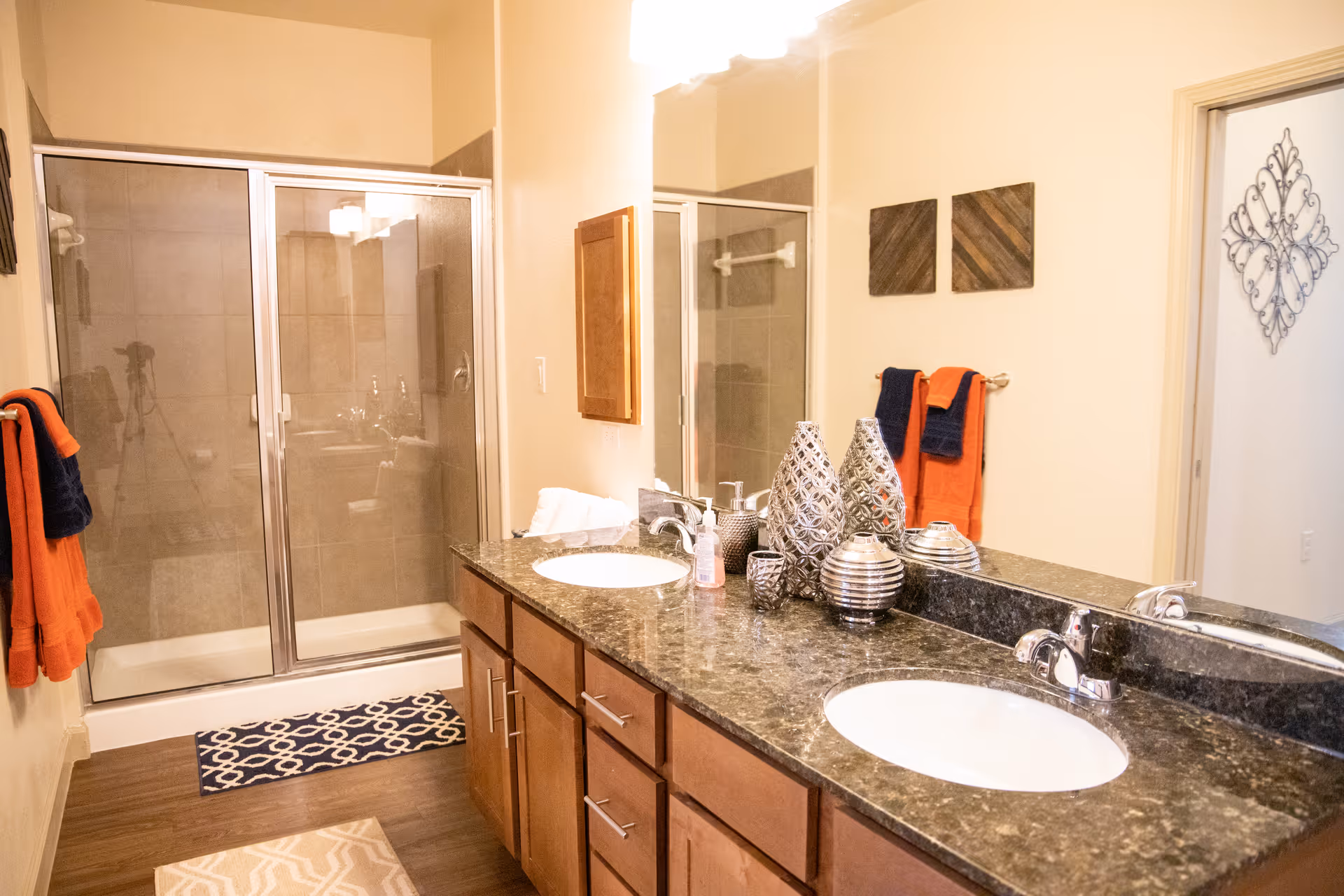 A bathroom with a double sink vanity featuring a dark granite countertop and wooden cabinets. There are decorative vases and a soap dispenser on the counter. The bathroom has a glass-enclosed shower with beige tiles. Two sets of towels, one orange and one dark blue, hang on the wall. The floor has two patterned rugs, and there is wall art and a decorative metal piece near the door.
