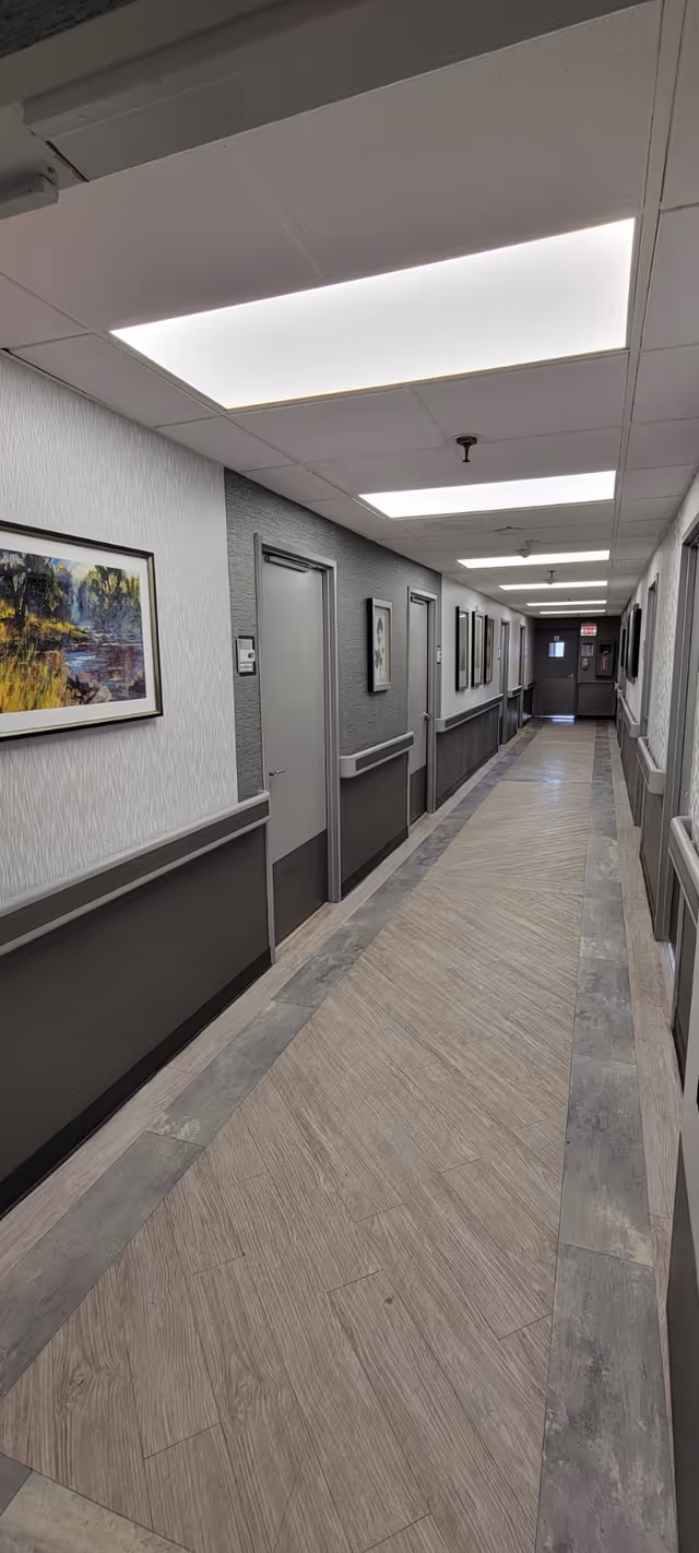 A long, well-lit hallway in a senior living facility with gray walls and doors on both sides. The floor has a wood-like pattern bordered by a gray tile design. Several framed paintings hang on the walls, and there are handrails along both sides of the hallway. The ceiling has recessed fluorescent lighting panels.