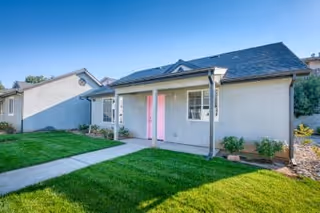Exterior view of a single-story residential building with a gray roof and light-colored walls, featuring a small covered porch with a pink door, surrounded by a well-maintained green lawn and a concrete walkway leading to the entrance.
