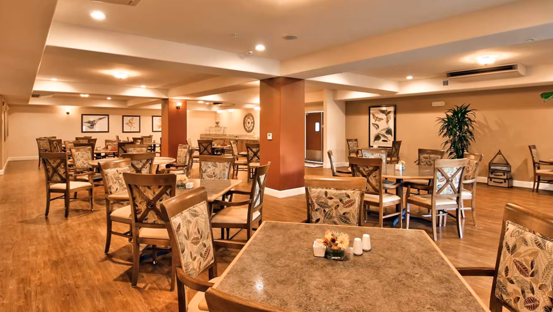 A spacious dining room with multiple wooden tables and chairs featuring leaf-patterned cushions. The room has warm lighting, wood flooring, and beige walls decorated with framed bird artwork and a large clock. There are plants and decorative items placed around the room.