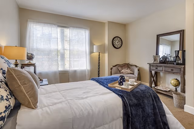 A cozy bedroom with a neatly made bed featuring white and blue bedding. There is a wooden side table with a lamp and a globe on it next to the bed. A window with sheer curtains allows natural light to fill the room. In the corner, there is a cushioned chair with a pillow, a floor lamp, and a wall clock above it. A wooden dresser with a mirror, decorative items, and a globe sits against the wall.