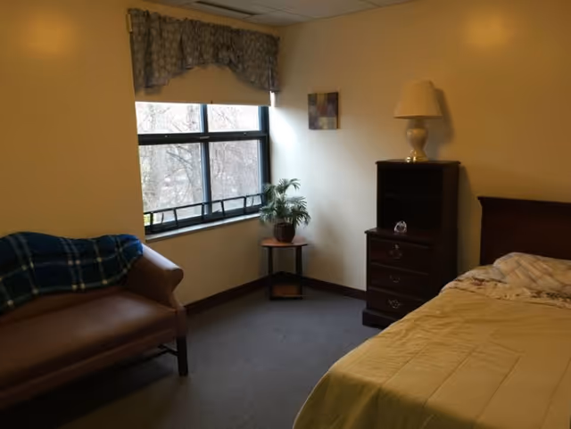 A simple bedroom with a bed covered in a yellow blanket, a wooden nightstand with a lamp, a small table with a potted plant, a window with a valance, and a brown couch with a blue plaid blanket draped over one arm.