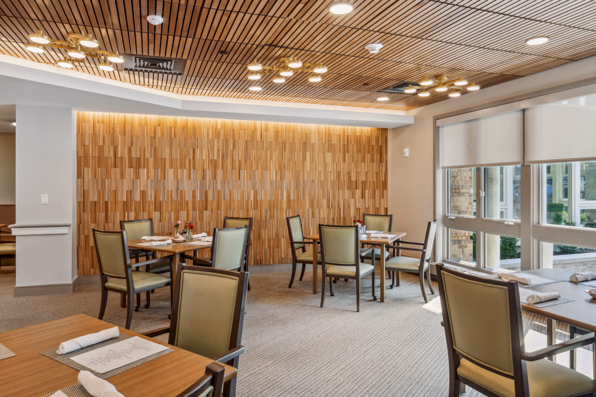 Bright dining room with several tables and chairs arranged neatly. The room features a wooden slatted ceiling with modern light fixtures, a textured wooden accent wall, large windows with roller shades allowing natural light, and tables set with placemats, napkins, and menus.