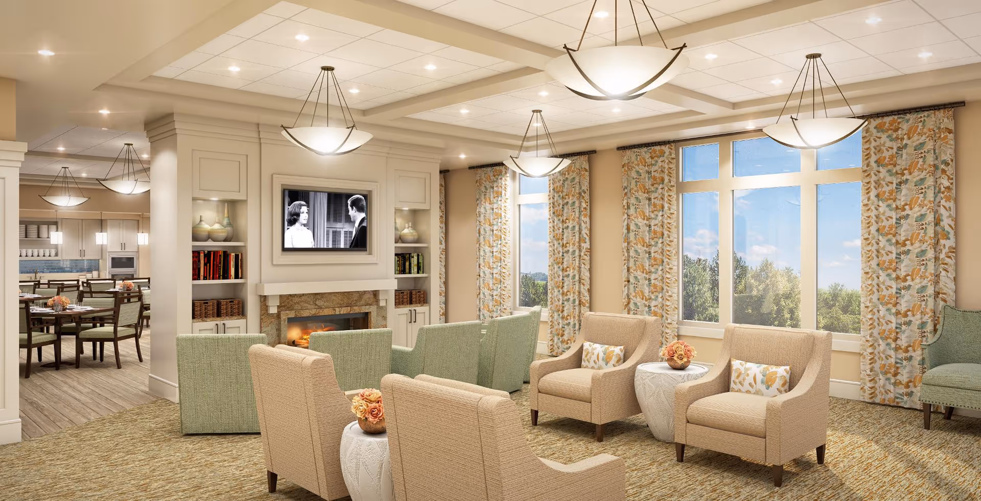 A bright and spacious living room area with beige and green upholstered chairs arranged around small round tables with floral centerpieces. The room features large windows with floral curtains letting in natural light, a fireplace with a built-in TV above it, and a view into an adjacent dining area with tables and chairs. The ceiling has recessed lighting and hanging light fixtures.
