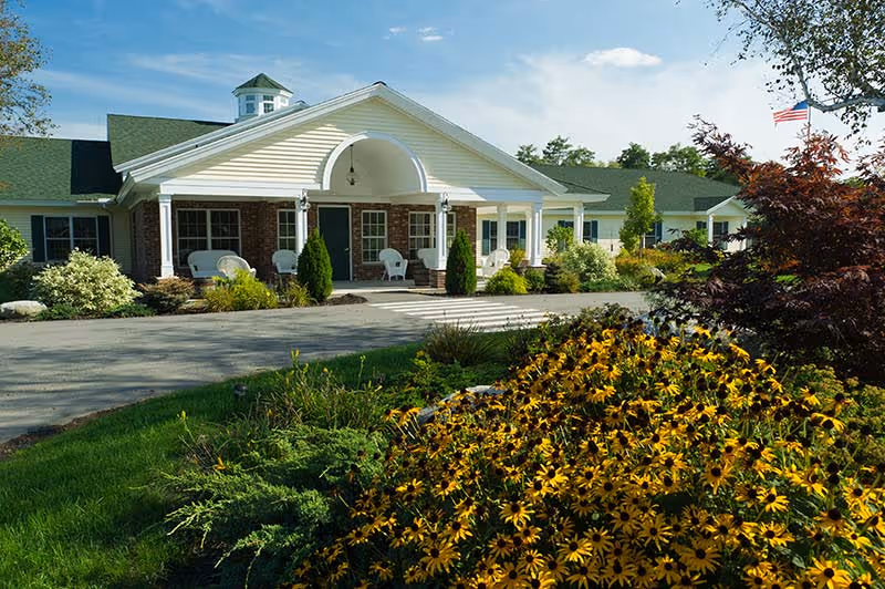 Front exterior view of a single-story senior living facility building with a covered entrance, white columns, and a well-maintained garden with yellow flowers and shrubs in the foreground under a blue sky.