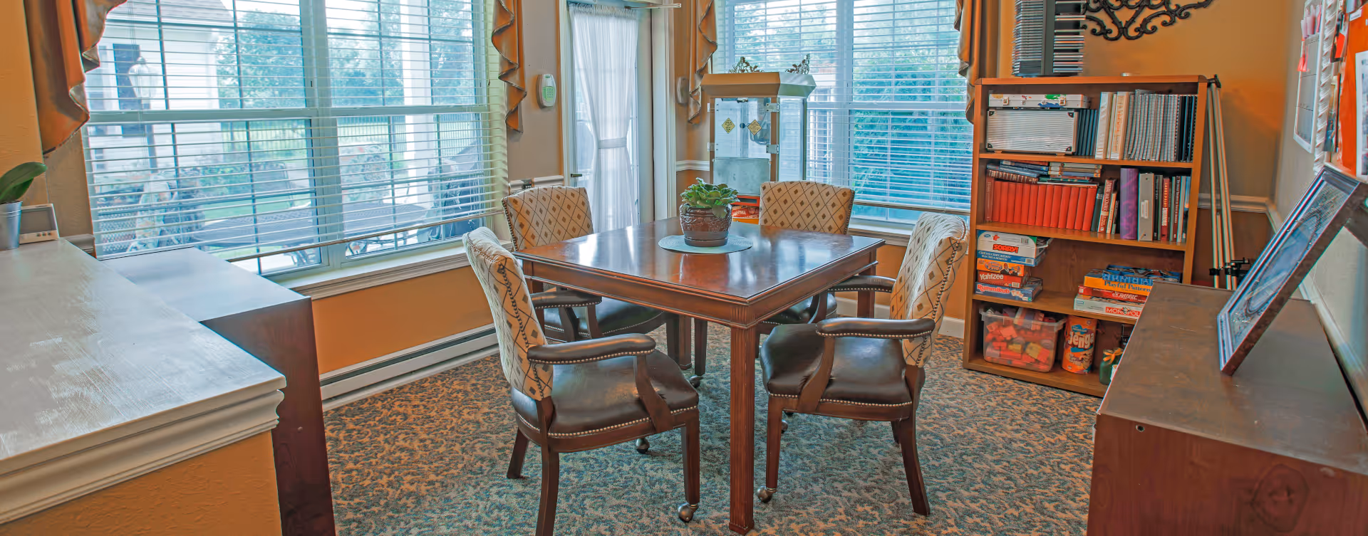 A cozy room with a square wooden table surrounded by four cushioned chairs with patterned upholstery. The room has large windows with blinds and orange curtains, letting in natural light. There is a bookshelf filled with books and board games, and a small plant is placed on the table. The carpet has a floral pattern, and the walls are painted a warm beige color.
