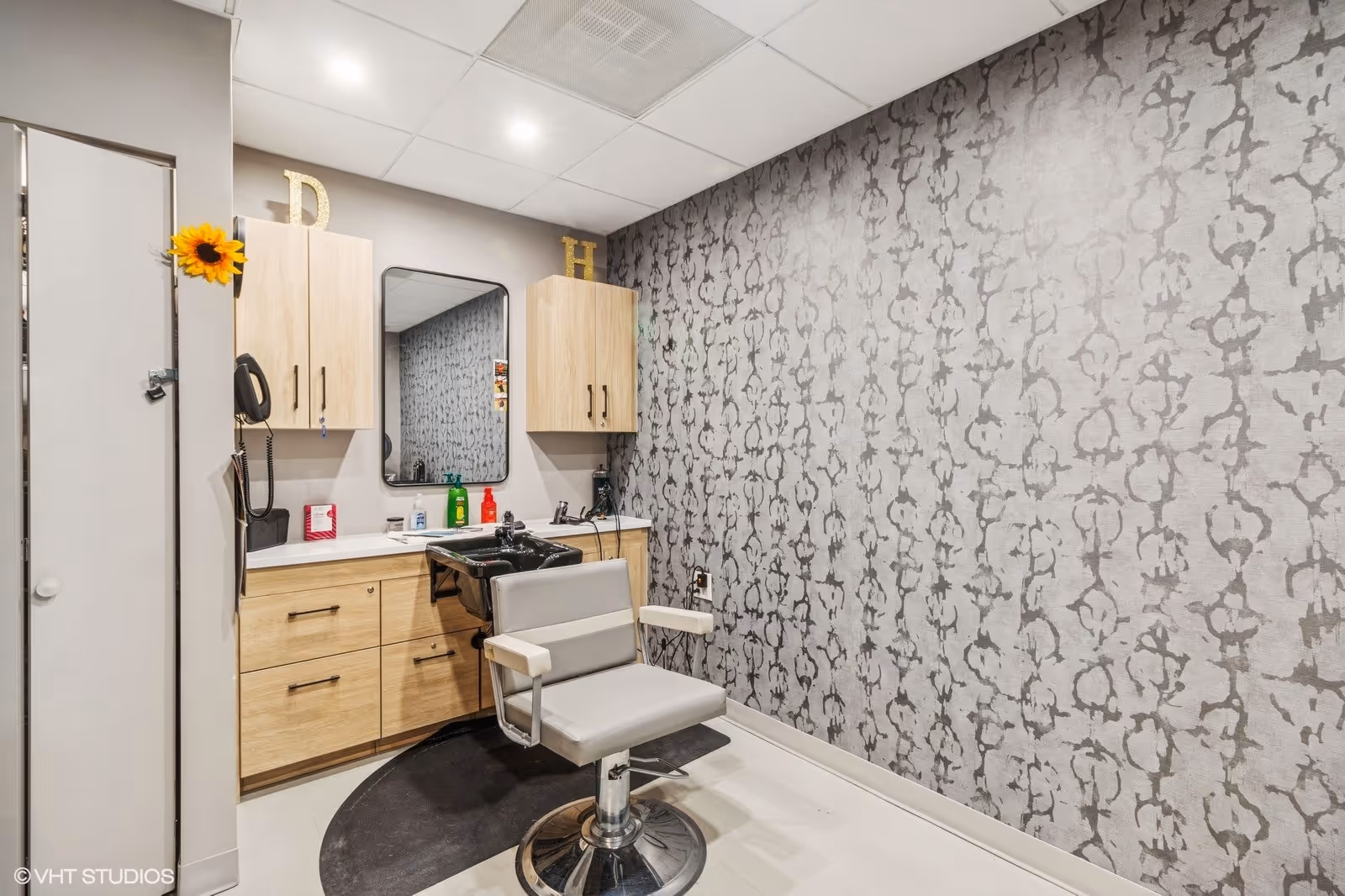 Interior view of a hair salon area in a senior living facility with a salon chair in front of a black sink, a mirror mounted on the wall, wooden cabinets, and patterned wallpaper on one side.