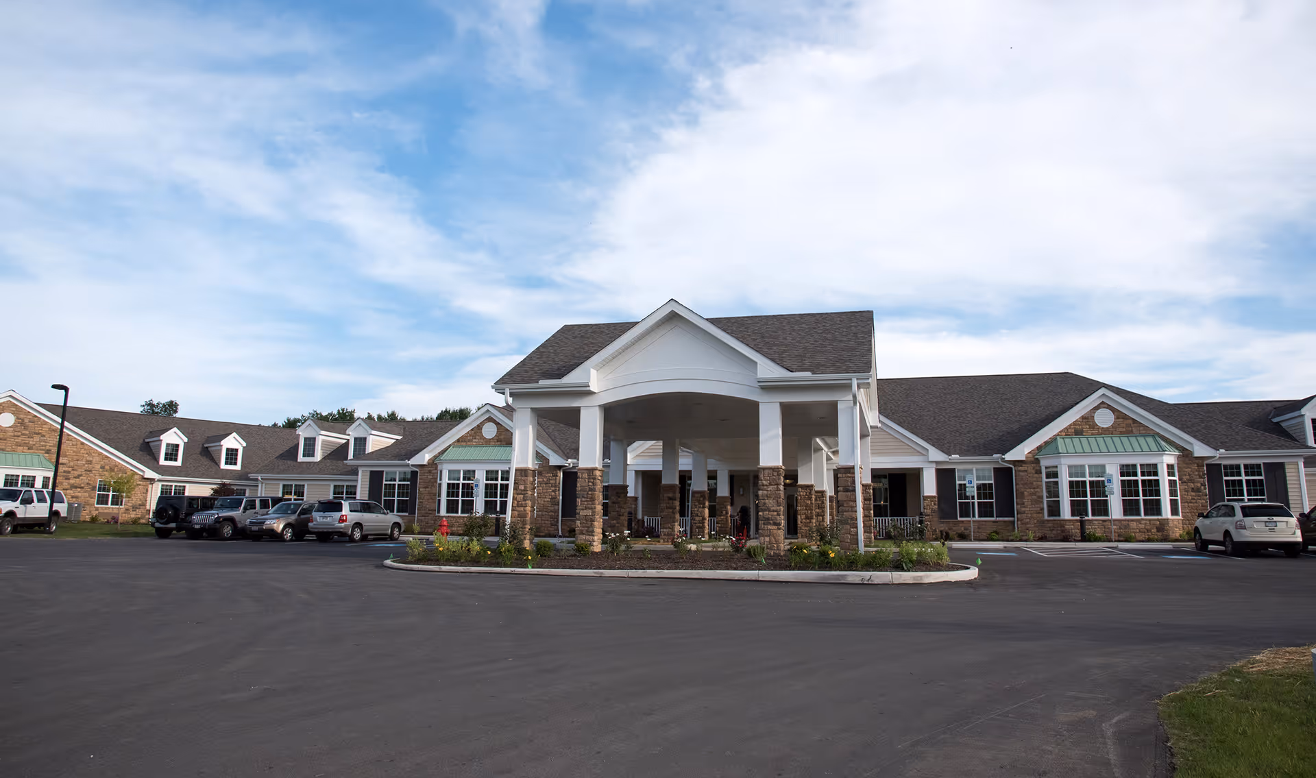 Front exterior view of Mapleview Country Villa, a single-story building with a covered entrance supported by white columns and stone bases, surrounded by a parking lot with several parked cars under a partly cloudy sky.