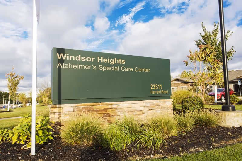Outdoor view of a green and stone sign for Windsor Heights Alzheimer's Special Care Center located at 23311 Harvard Road, surrounded by landscaping with grass, bushes, and trees under a partly cloudy sky.