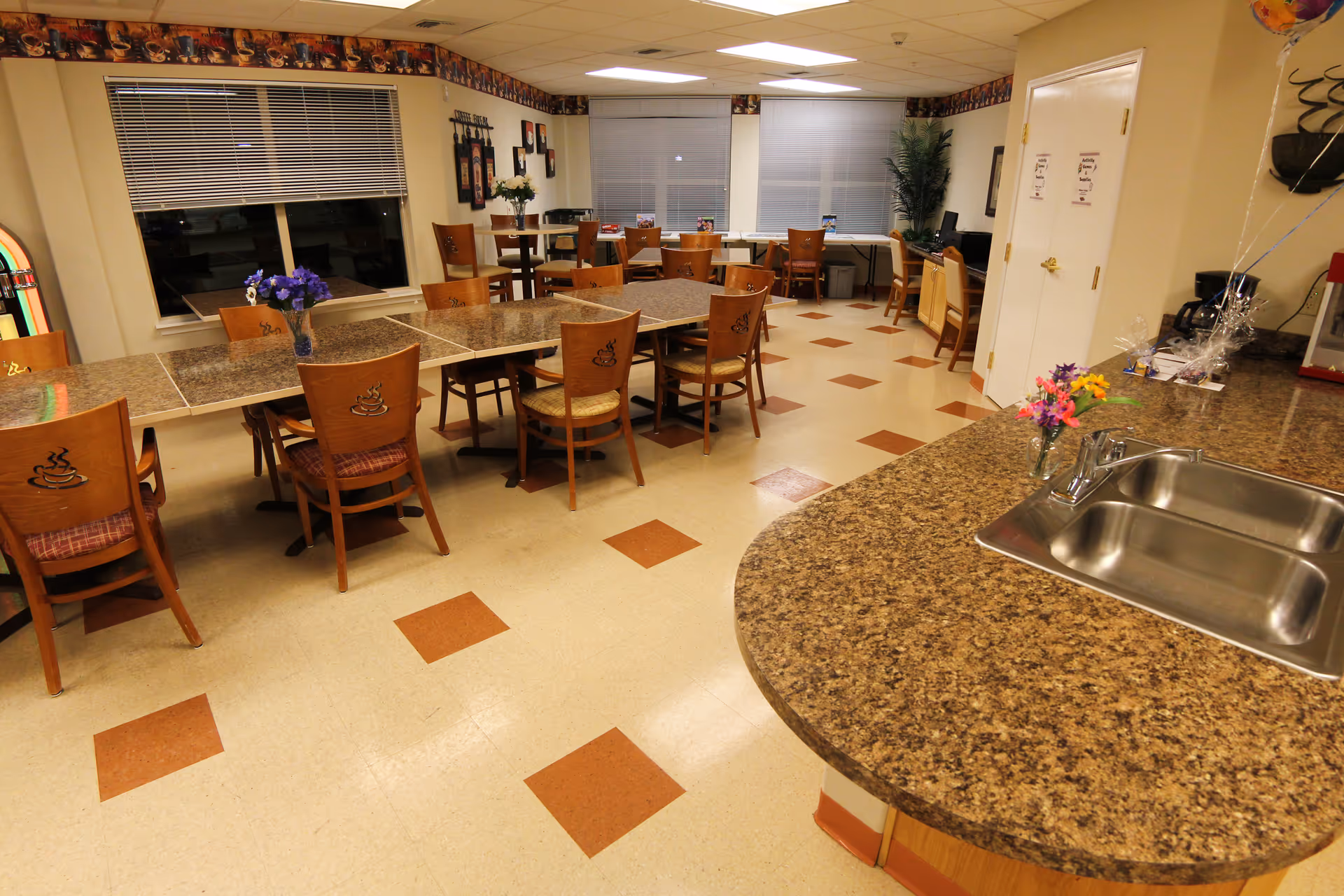 Interior view of a dining room in a senior living facility with multiple wooden tables and chairs arranged neatly. The tables have a brown speckled surface, and some have small flower vases on them. There is a countertop with a stainless steel sink in the foreground on the right side. The room has beige walls, a tiled floor with brown square accents, and windows with blinds. Decorations and a plant are visible in the background.