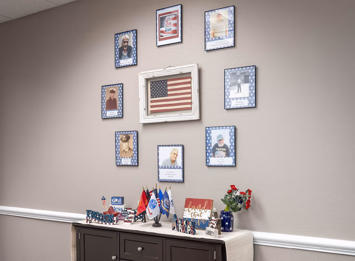 A beige wall decorated with framed photos of veterans surrounding a framed American flag. Below the photos is a dark wooden cabinet with patriotic decorations including small flags, a sign that says 'land that I love', and a vase with red flowers.