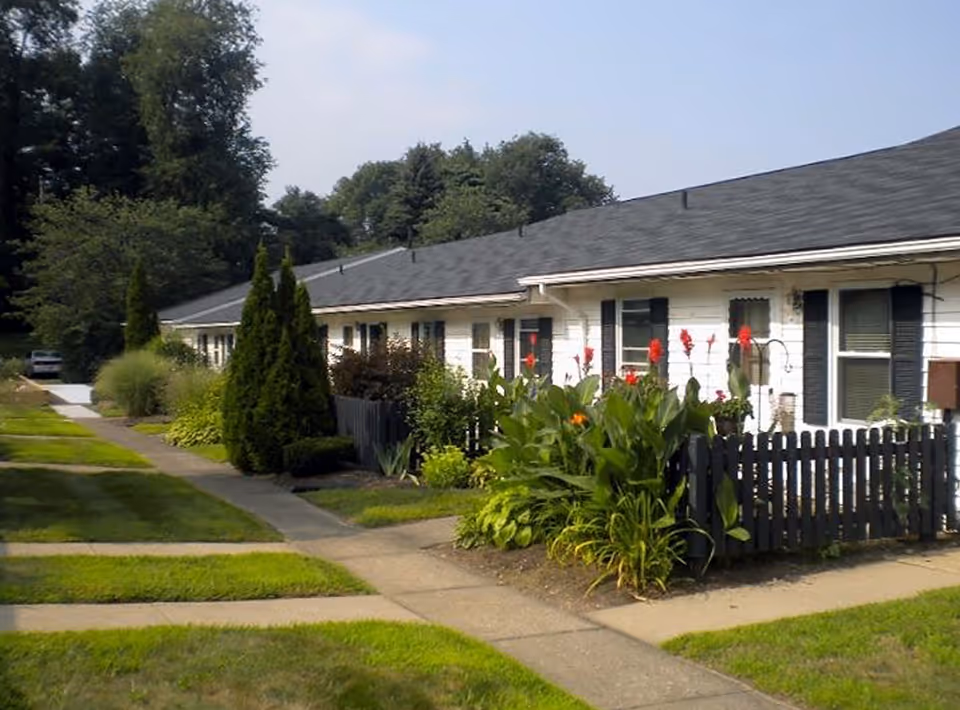 Exterior view of a single-story residential building with white siding and black shutters. The building is surrounded by well-maintained green lawns, bushes, and flowering plants. A concrete walkway runs alongside the building, and there are trees in the background under a clear sky.