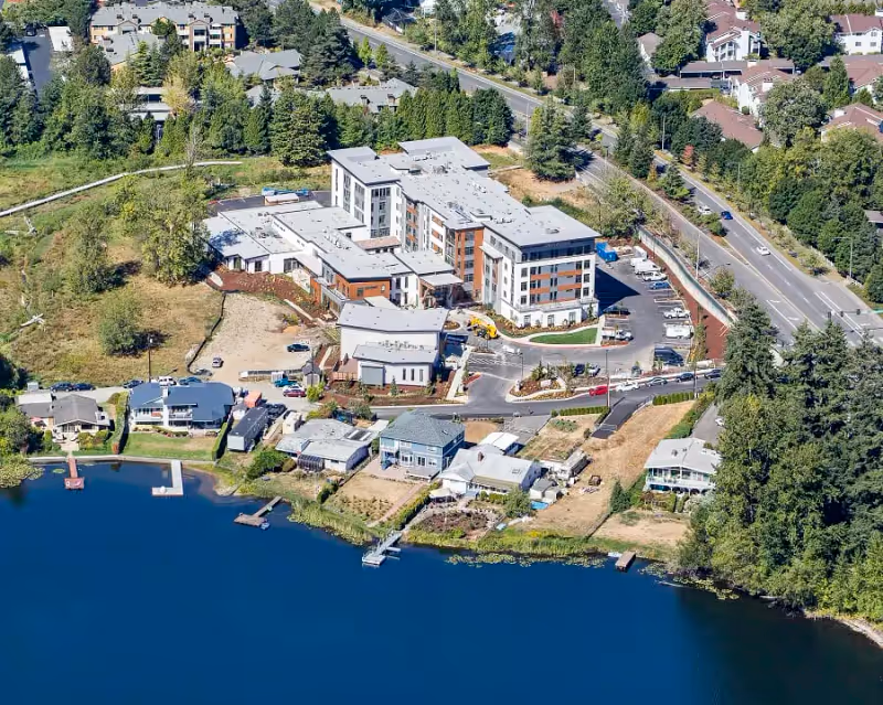 Aerial view of Aegis Gardens Newcastle, a multi-story senior living facility surrounded by trees, roads, and residential houses near a large body of water with docks.