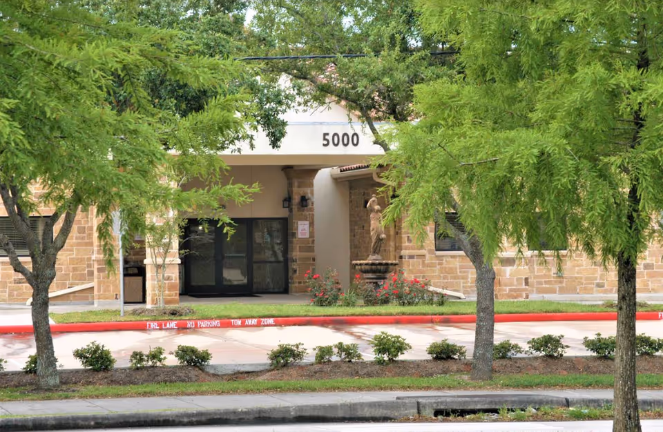 Entrance of a building with the number 5000 above the doorway, surrounded by green trees and shrubs. There is a stone statue fountain near the entrance and a red curb with 'FIRE LANE NO PARKING TOW AWAY ZONE' painted on it.