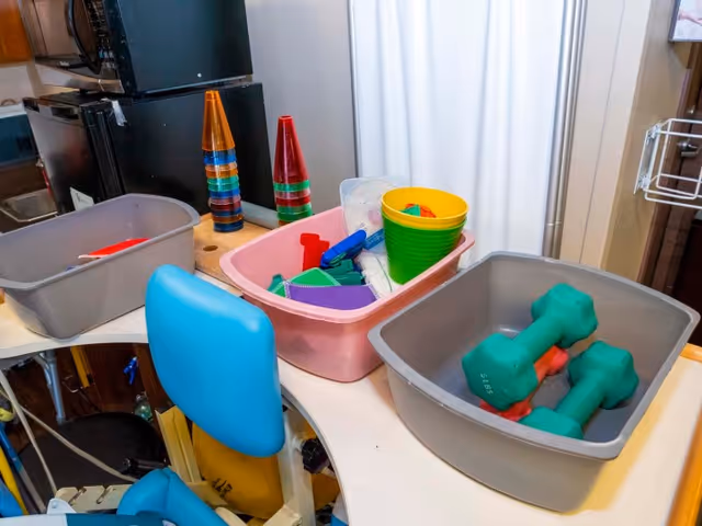 A therapy or activity room with a curved white table holding three plastic bins. One bin contains green and orange dumbbells, another has colorful plastic cones and small toys, and the third has various small items including a green cup. Blue padded chairs are positioned around the table, and a black refrigerator and microwave are visible in the background.