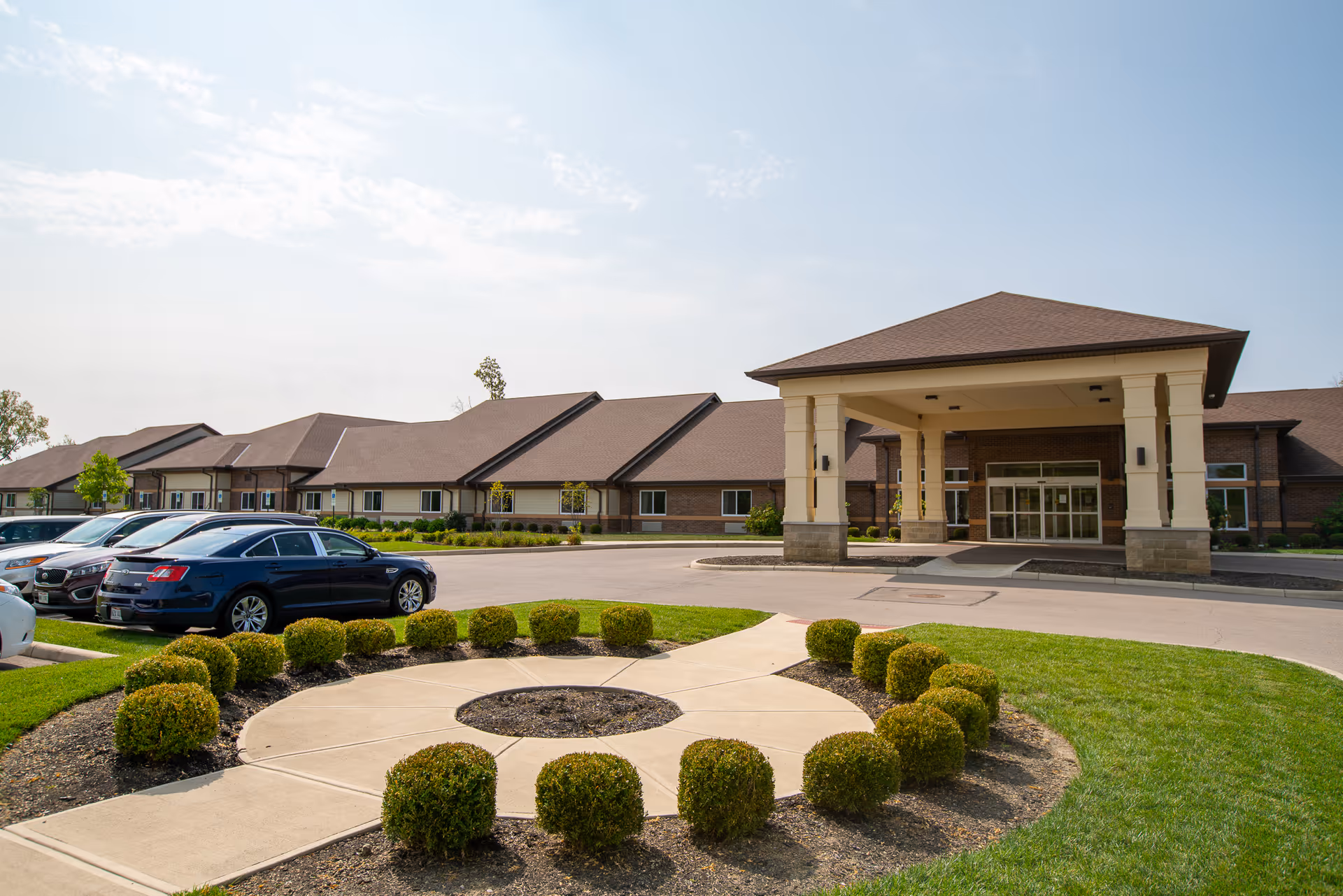 Exterior view of Austin Trace Health and Rehabilitation facility showing a large covered entrance with pillars, a circular landscaped area with trimmed bushes, a parking lot with several cars, and a clear sky.