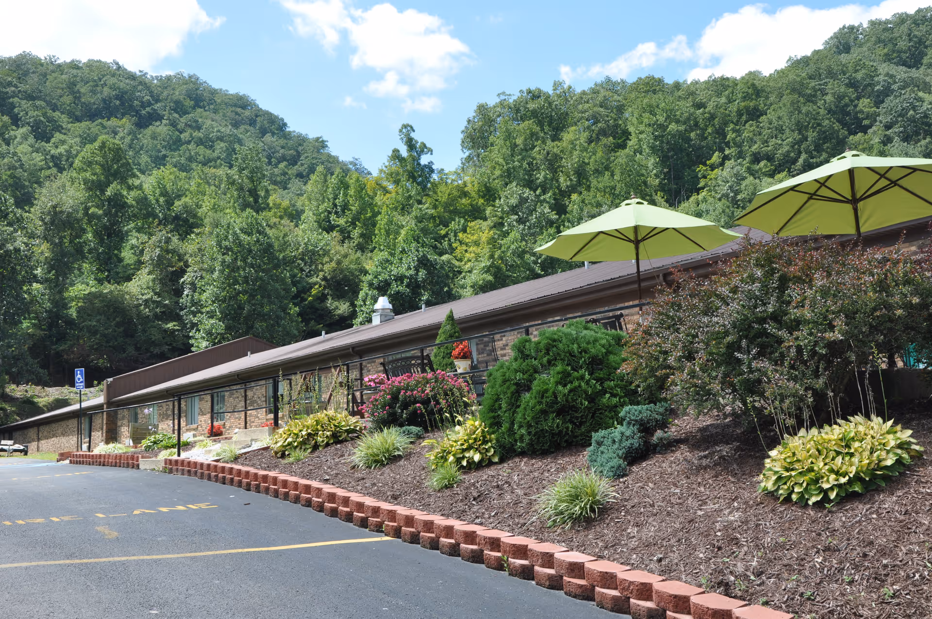 Exterior view of Good Shepherd Health and Rehabilitation Center showing a single-story brick building with a landscaped garden in front, including bushes, flowers, and two green patio umbrellas. The building is surrounded by lush green trees and hills under a partly cloudy sky. A paved parking area with a handicapped parking sign is visible in the foreground.