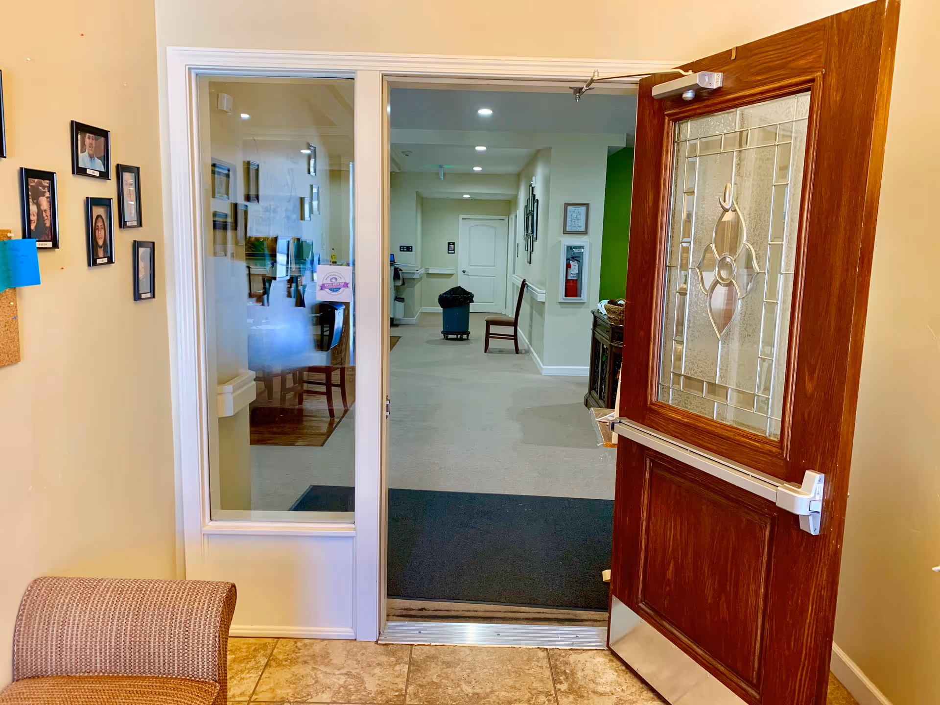 View through an open wooden door with decorative glass panel into a hallway of a senior living facility. The hallway has beige walls, carpeted floor, framed pictures on the walls, a fire extinguisher, and a chair. To the left of the door, there is a wall with several framed photos and a corkboard with a blue sticky note. A cushioned chair is partially visible in the foreground.