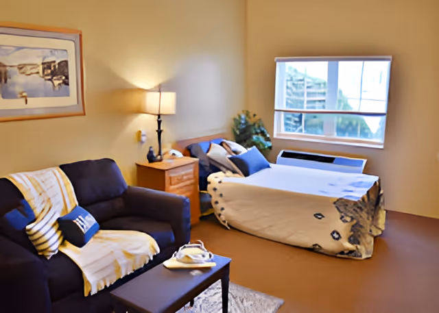 A cozy bedroom in a senior living facility featuring a bed with a patterned blanket, a nightstand with a lamp, a small sofa with cushions and a throw blanket, a coffee table with a tray, a framed picture on the wall, a window with a view outside, and a potted plant in the corner.