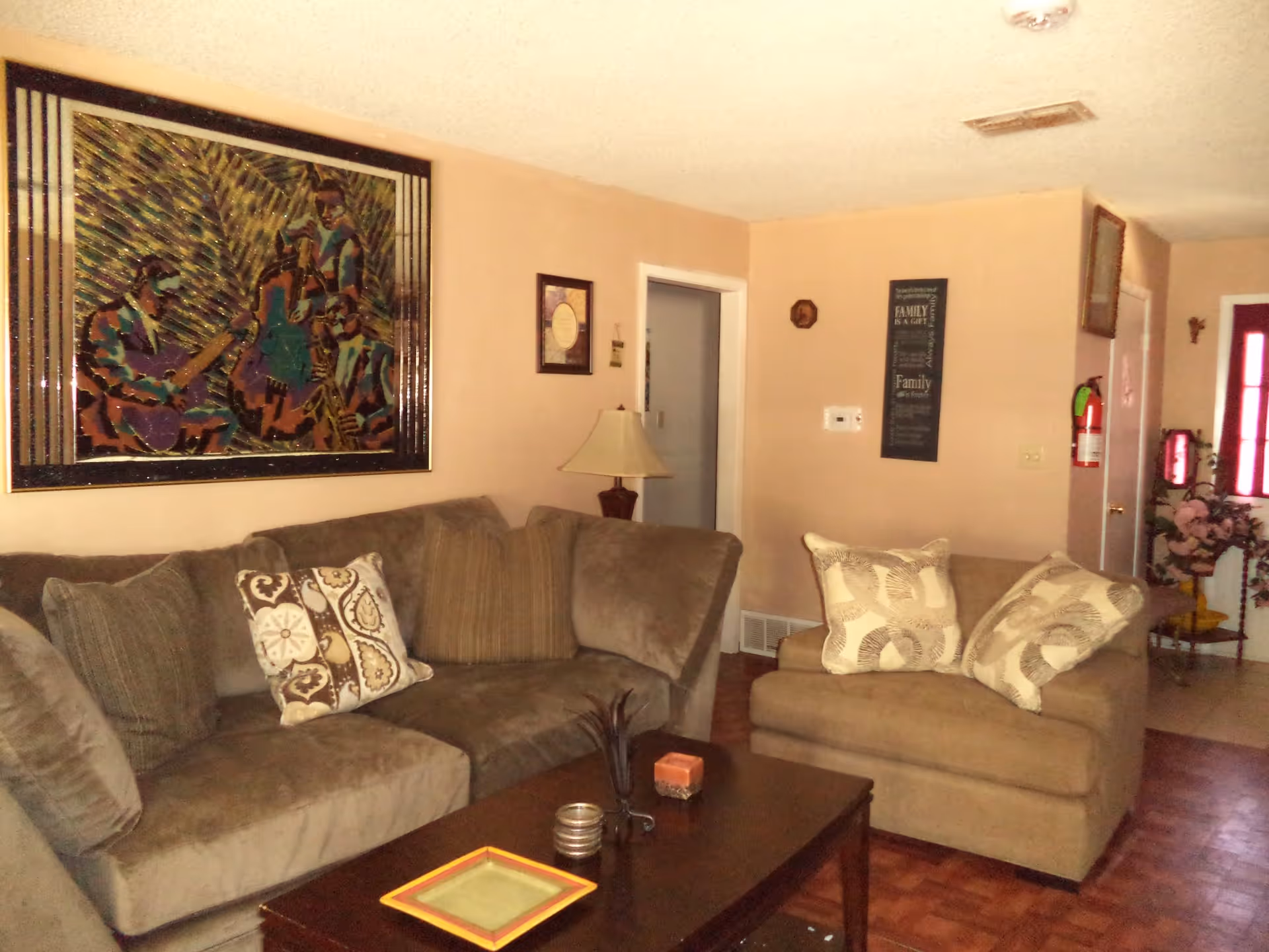 A cozy living room with a brown sectional sofa and a matching armchair, both adorned with patterned throw pillows. A dark wooden coffee table sits in front of the sofa with decorative items on it. The walls are painted beige and decorated with framed artwork and a lamp on a side table. A fire extinguisher is mounted near the entrance door in the background.
