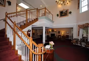 Interior view of a senior living facility featuring a carpeted staircase with wooden handrails leading to an upper floor. The area includes a sitting space with chairs, a chandelier hanging from the ceiling, framed artwork on the walls, and large windows allowing natural light.