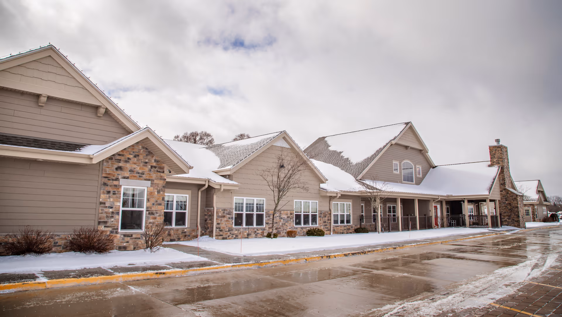 Exterior view of a large residential building with beige siding and stone accents, partially covered with snow on the roof and ground. The sky is cloudy and the driveway in front is wet with patches of snow.