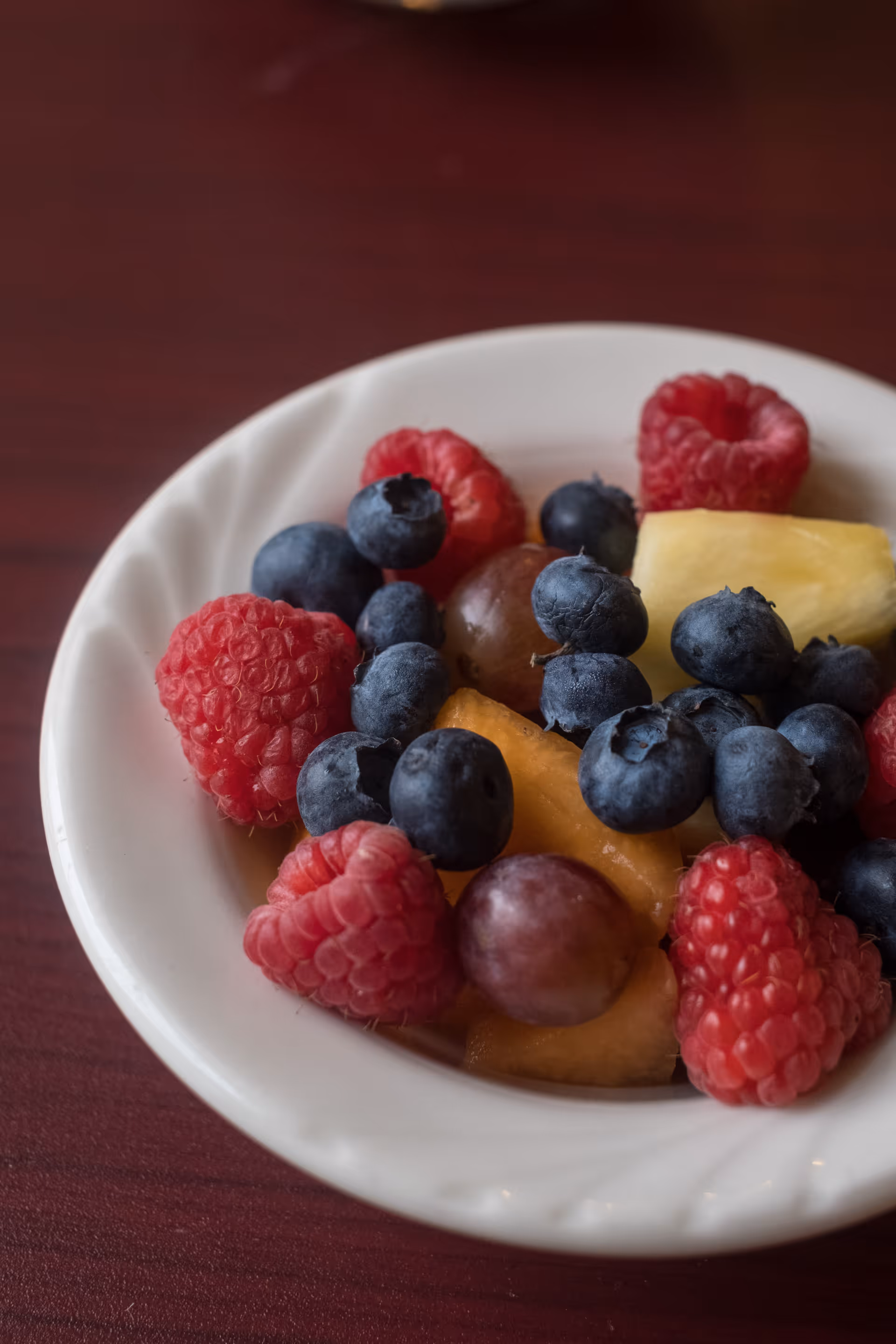 A white bowl filled with fresh mixed fruit including raspberries, blueberries, grapes, pineapple, and cantaloupe on a dark wooden table.