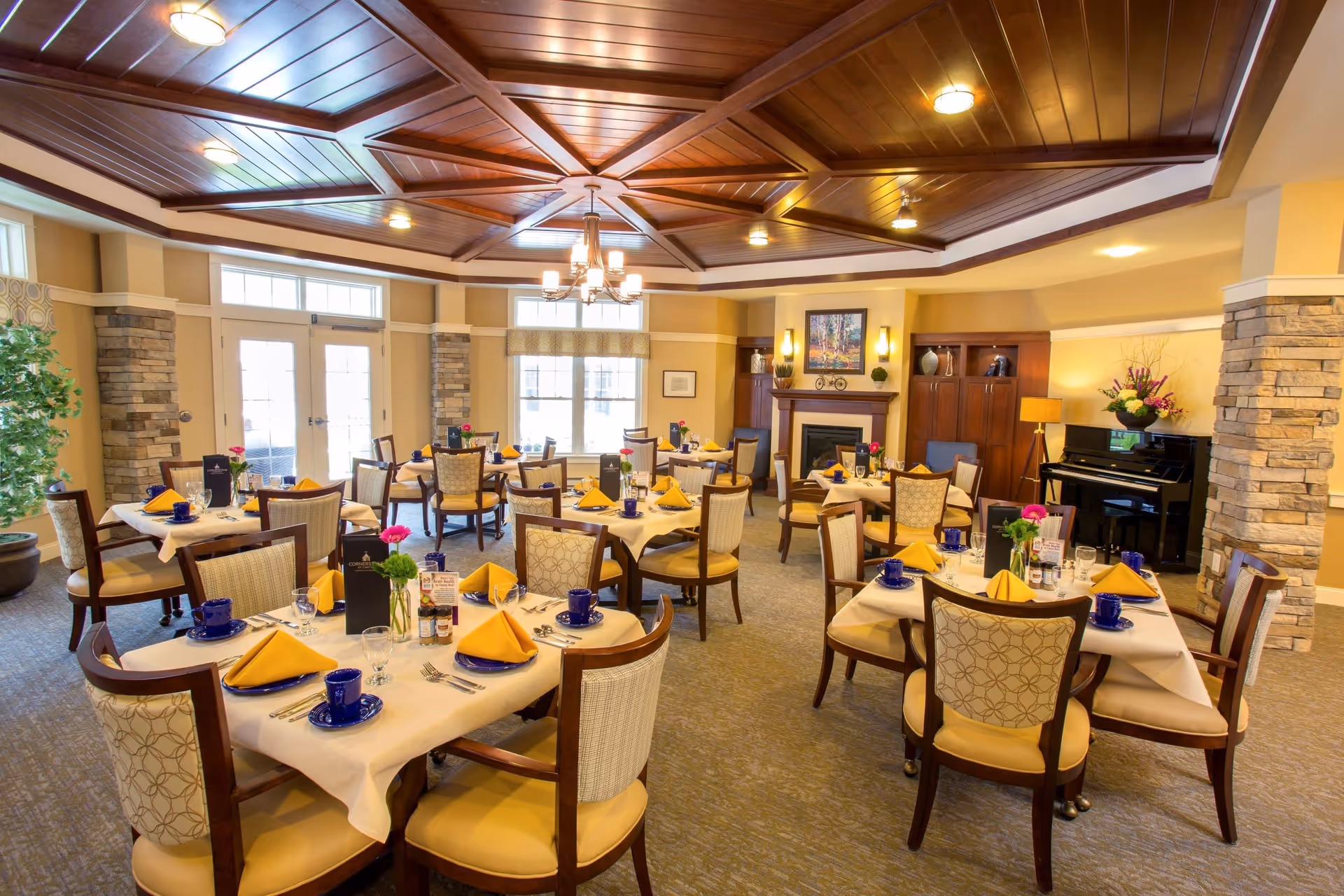 A bright and elegant dining room with multiple tables set with white tablecloths, yellow napkins, blue cups and saucers, and silverware. The room features a wooden coffered ceiling with a chandelier, stone pillars, large windows, a fireplace with a painting above it, and a black piano in the corner. There are decorative plants and flowers on the tables and around the room.