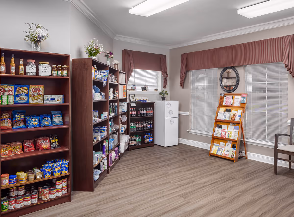 Interior view of a small convenience area in a senior living facility with wooden shelves stocked with snacks, canned goods, and personal care items. There is a small refrigerator and a display rack with brochures or cards near two windows with blinds and maroon valances. The floor is wood-patterned, and the walls are light-colored with crown molding.