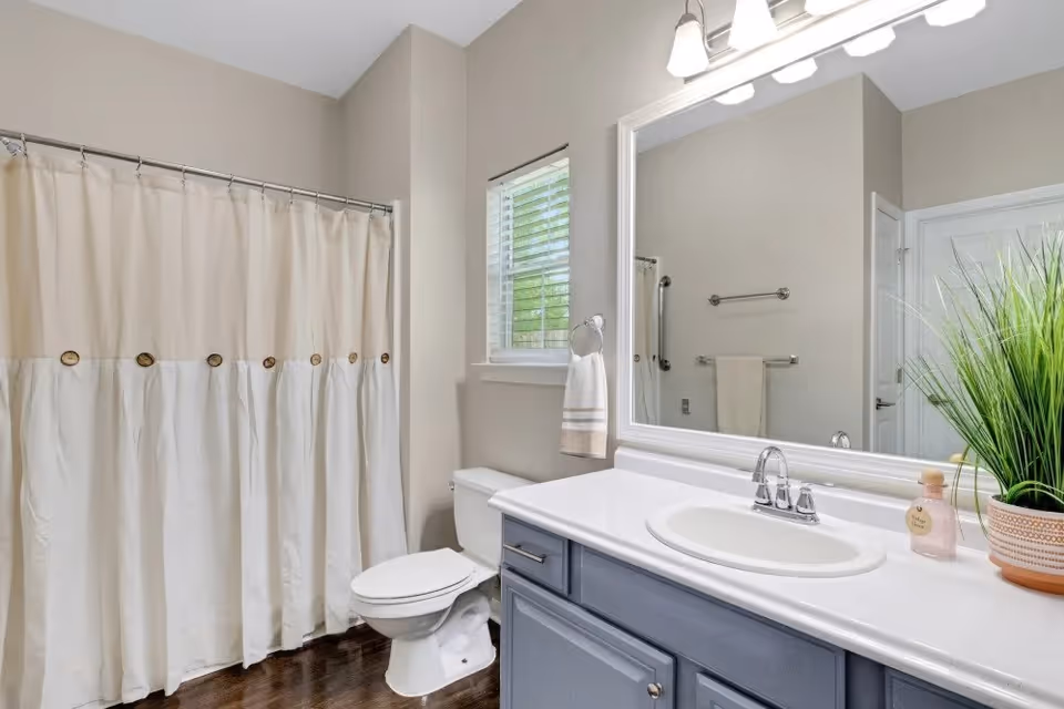 A clean and modern bathroom featuring a white toilet, a large mirror above a white sink with a blue vanity cabinet, a beige and white shower curtain, a small window with blinds, a towel hanging on a ring, and a potted plant on the countertop.