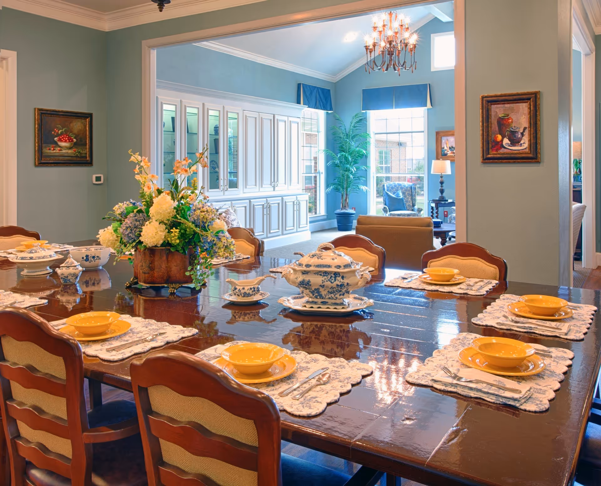 A formal dining room with a large polished wooden table set with yellow bowls, plates, and silverware on white lace placemats. A floral centerpiece decorates the middle of the table. The room has light blue walls with framed paintings, and an adjoining living area with large windows, a chandelier, and a potted plant.