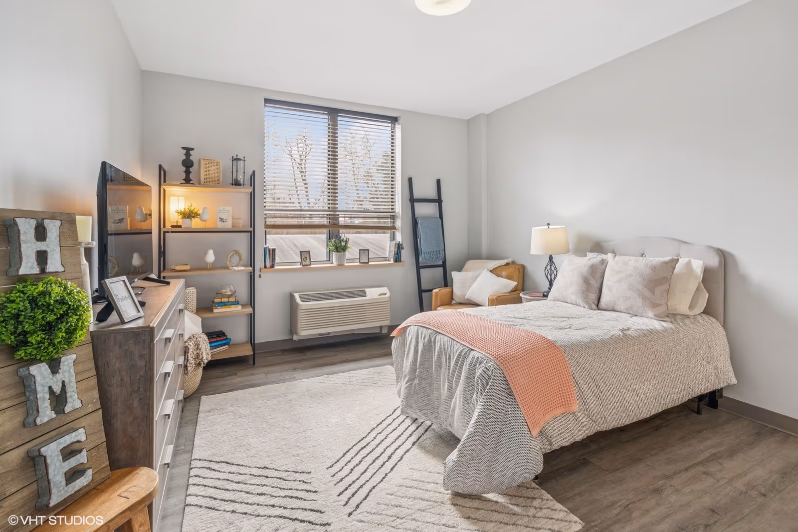 A bright and cozy bedroom with a large window featuring horizontal blinds. The room has a bed with light gray bedding and a coral throw blanket, a wooden dresser with a TV on top, a black metal shelving unit with decorative items, a beige armchair with a pillow, a floor lamp, and a decorative ladder with a blanket. The floor is wood with a light-colored area rug, and there is a wooden 'HOME' sign with greenery on the left side.