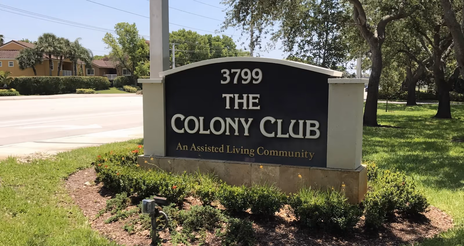 Outdoor view of a sign for The Colony Club, an assisted living community, located at 3799. The sign is surrounded by greenery and trees, with a street and residential buildings visible in the background.
