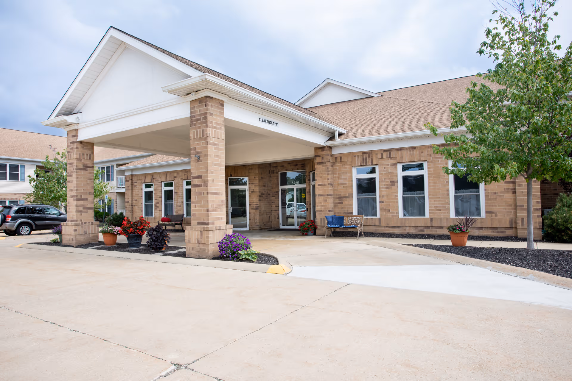 Exterior view of Governor's Port facility entrance with a covered drop-off area supported by brick columns, potted plants, benches, and a tree on the right side under a cloudy sky.