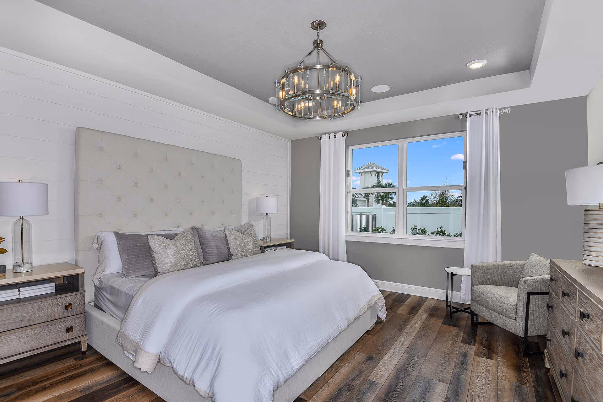 A bright and modern bedroom featuring a large bed with a tufted headboard, white bedding, and multiple pillows. The room has wooden flooring, two bedside tables with lamps, a comfortable armchair, a dresser, and a window with white curtains showing an outdoor view with a fence and greenery. A stylish chandelier hangs from the ceiling.
