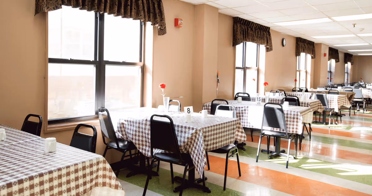 A bright dining room with multiple tables covered in checkered tablecloths. Each table has black chairs and a small vase with a red flower. Large windows with dark curtains allow natural light to fill the room. The floor has a colorful pattern with green, orange, and white stripes.