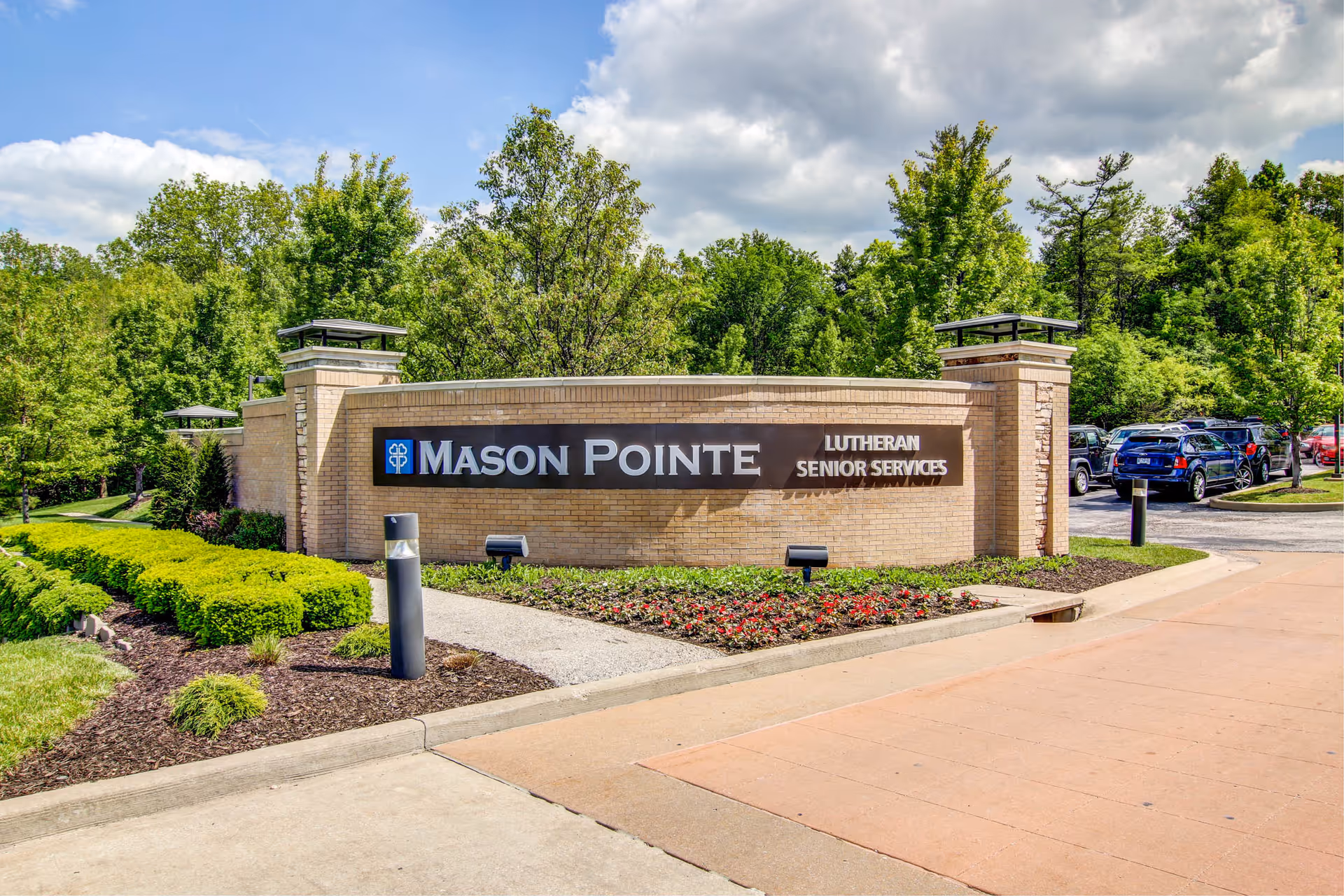 Entrance sign for Mason Pointe Lutheran Senior Services surrounded by landscaped greenery and trees, with a parking area and cars visible in the background under a partly cloudy sky.