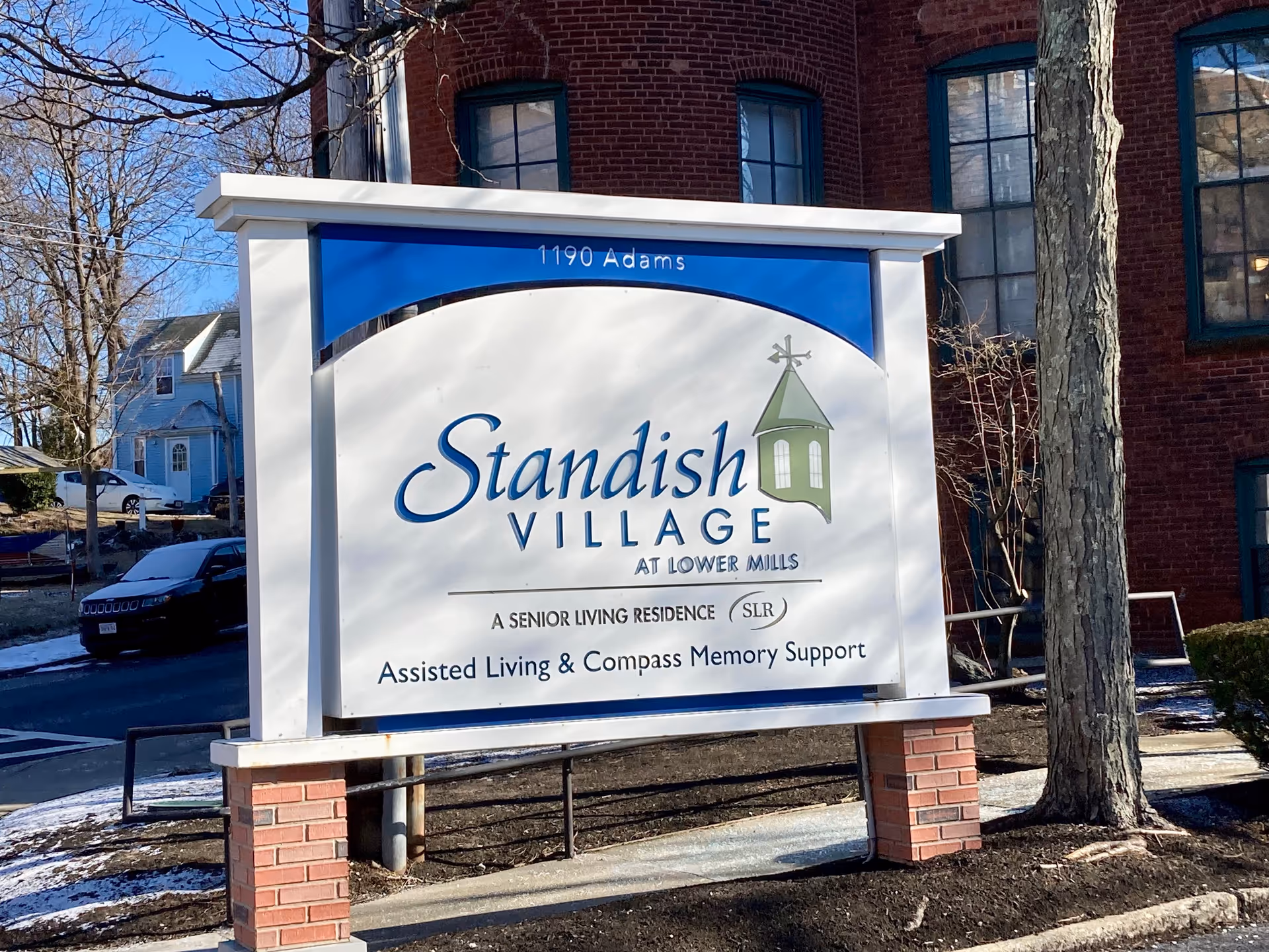 Outdoor sign for Standish Village at Lower Mills, a senior living residence offering Assisted Living and Compass Memory Support, located at 1190 Adams. The sign is white with blue and green accents, set against a backdrop of a brick building and trees.