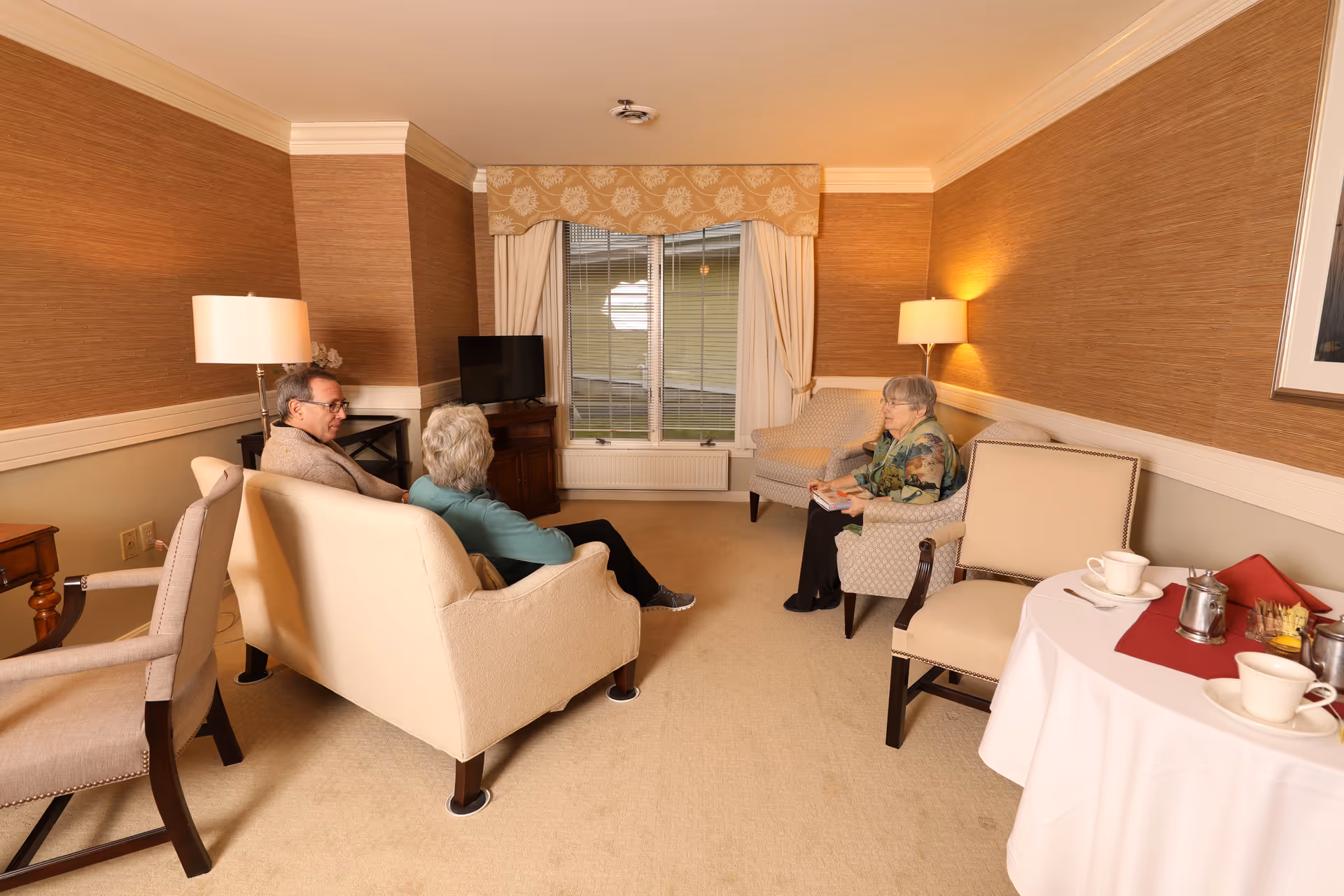 A cozy living room with beige walls and carpet, featuring three elderly people sitting and conversing. Two people sit on a beige loveseat facing a woman seated in an armchair. There is a small TV on a wooden stand near a window with cream-colored curtains and a valance. A round table with a white tablecloth holds cups, saucers, and a silver teapot. The room is softly lit by two floor lamps.