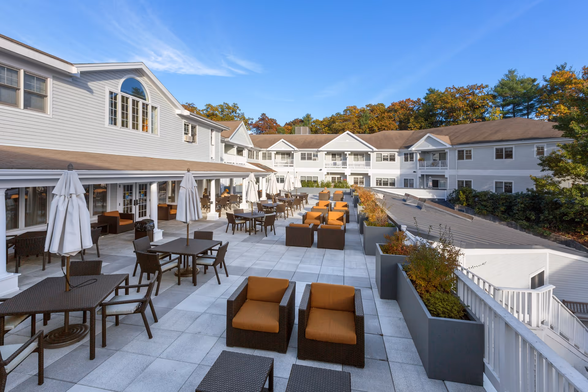 Outdoor patio area at Maplewood at Weston with multiple tables and chairs, some with white umbrellas, and cushioned lounge chairs arranged in rows. The patio is surrounded by a two-story building with white siding and large windows, under a clear blue sky with trees in the background.