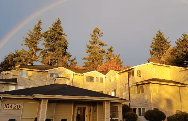 Exterior view of a senior living facility building with multiple windows and a covered entrance. Tall evergreen trees are visible behind the building, and a rainbow arcs across the sky above.