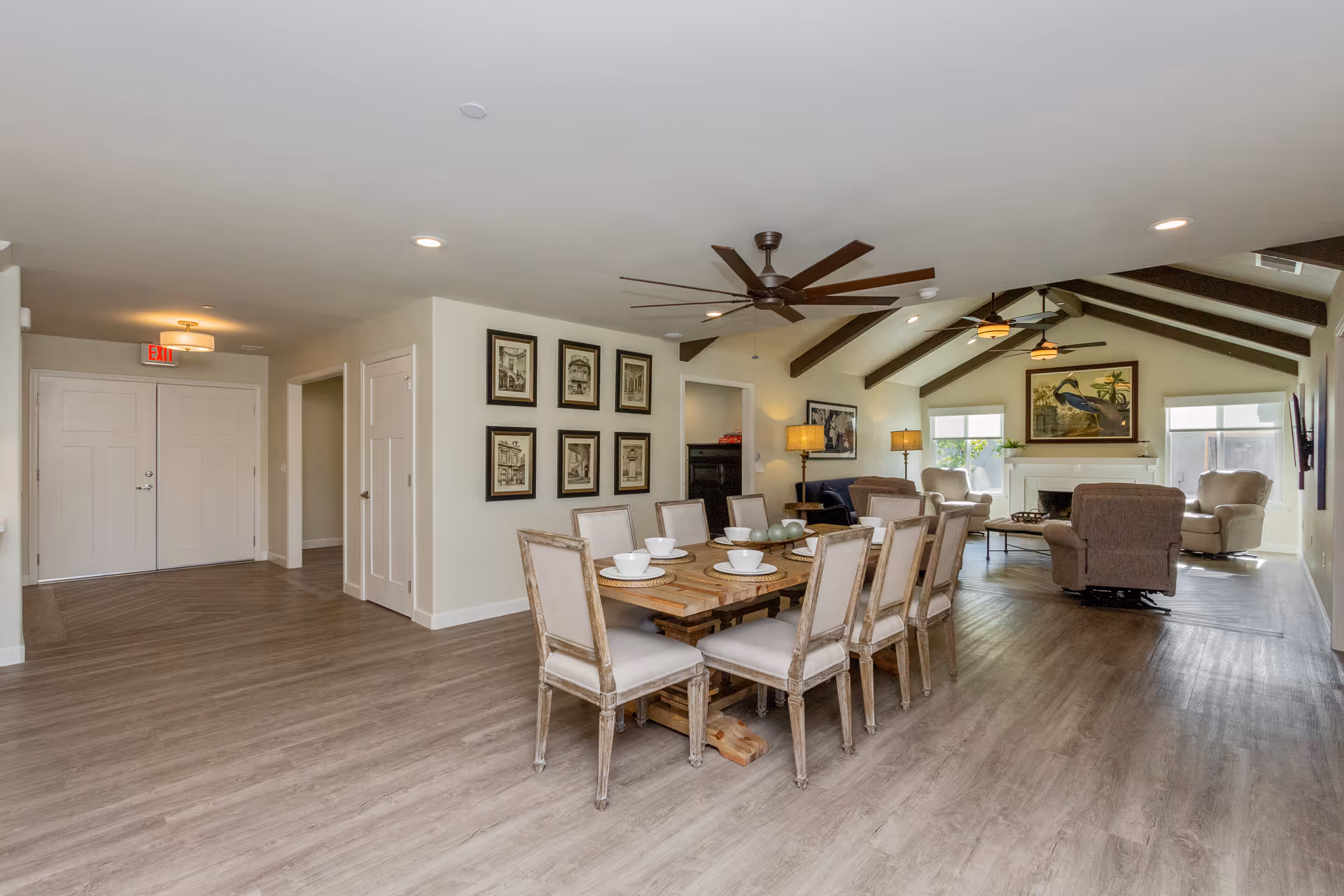 A spacious and well-lit common area in an assisted living facility featuring a long wooden dining table with eight chairs set with white bowls and plates. The room has light-colored walls, wood flooring, and ceiling fans with dark blades. In the background, there is a cozy living area with armchairs, a fireplace, framed artwork on the walls, and large windows letting in natural light.