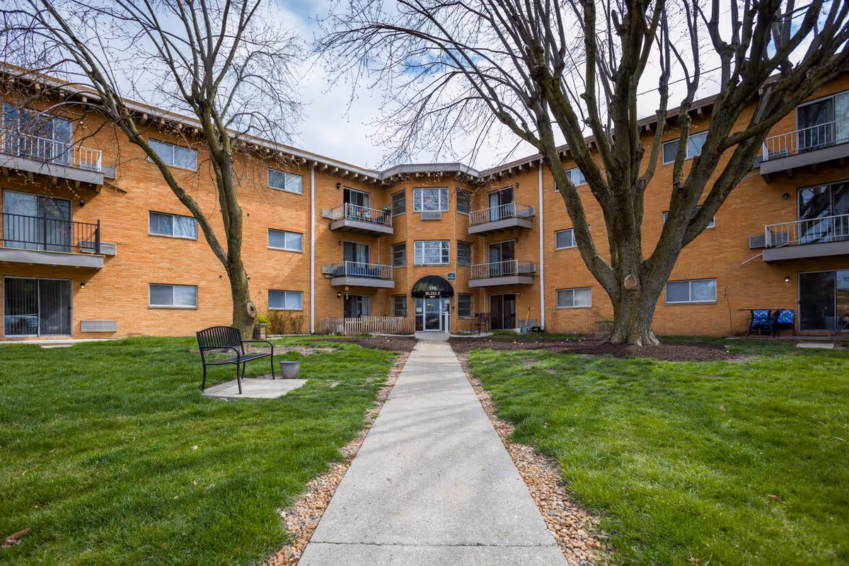 Exterior view of a three-story brick apartment building with balconies and large trees in front. A concrete walkway leads to the main entrance labeled 'Bldg B'. There is green grass and a bench near the walkway.